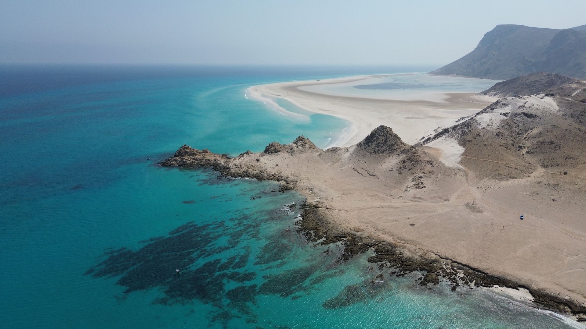 Aerial view of Socotra Island’s coastline with turquoise waters, white dunes, and dragon blood trees under a clear sky.