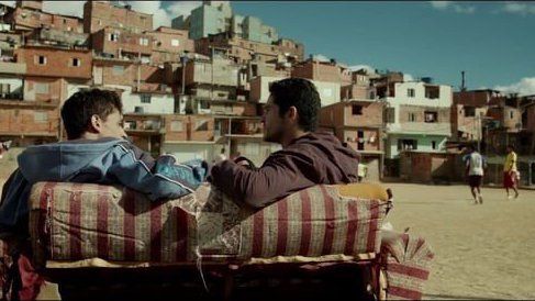 Dois homens estão sentados num banco em frente a um campo de futebol.