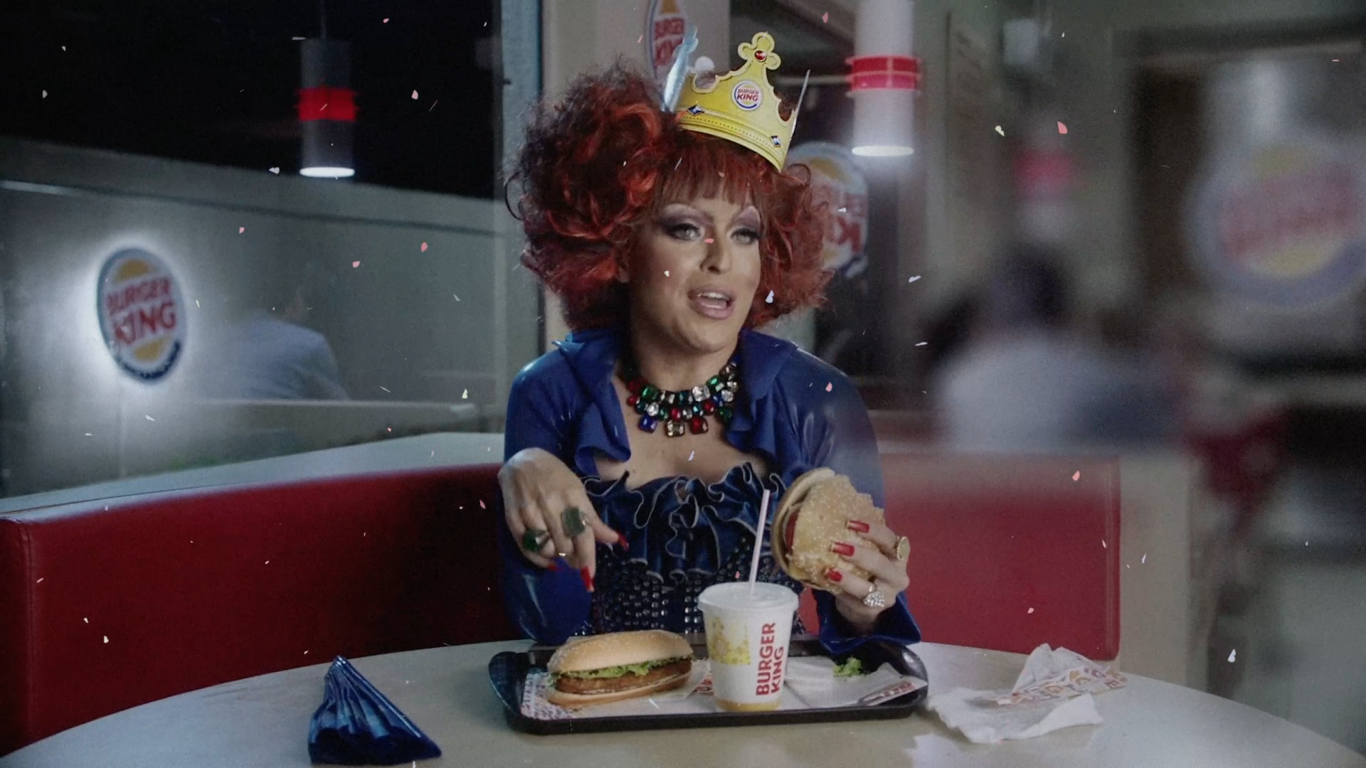 Uma drag queen está sentada à mesa de um restaurante Burger King comendo um hambúrguer.