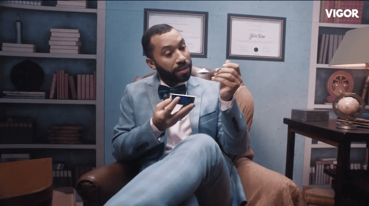 Um homem de terno e gravata borboleta está sentado em uma cadeira segurando um telefone celular.