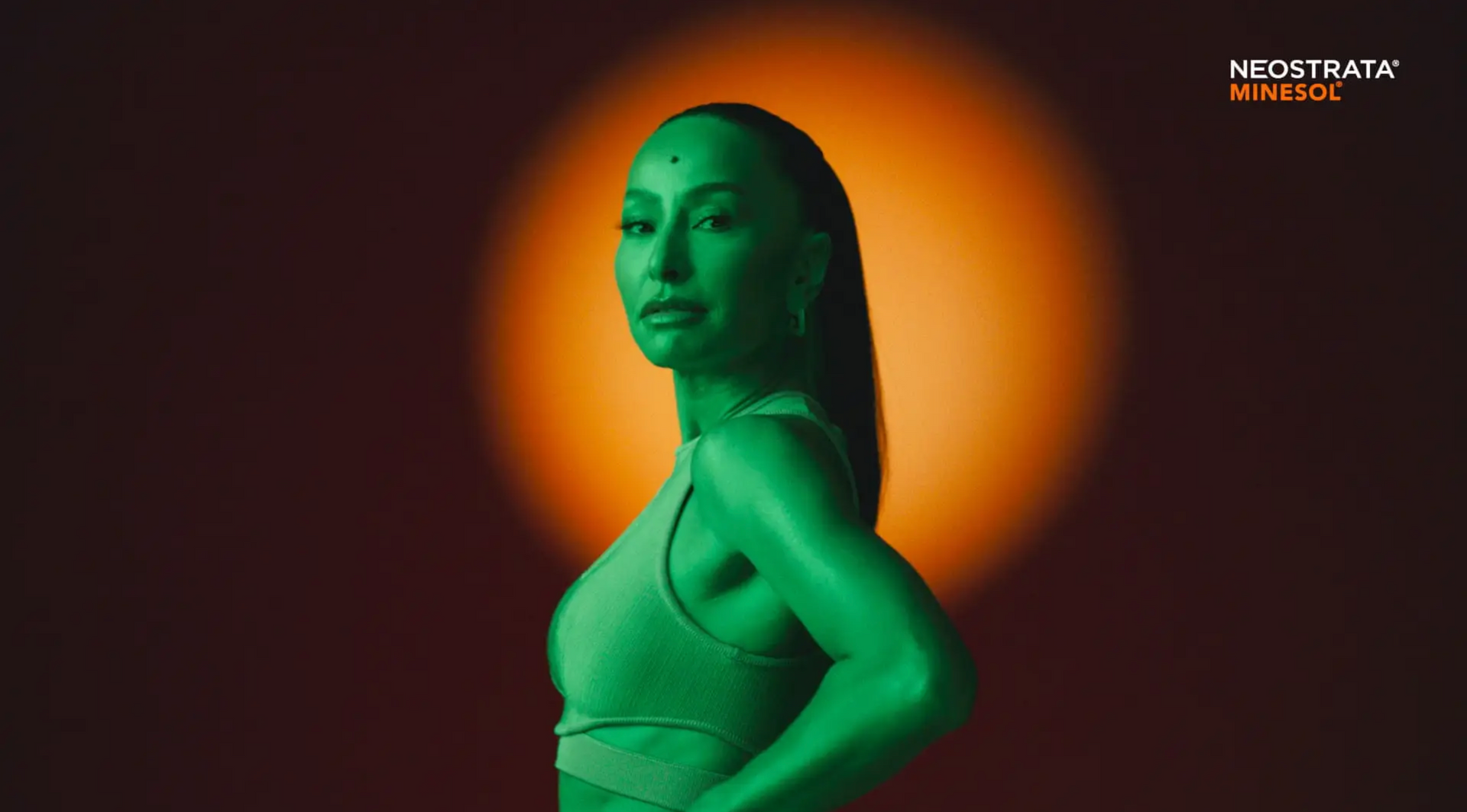 Uma mulher com uma blusa verde está parada em frente a uma luz laranja.