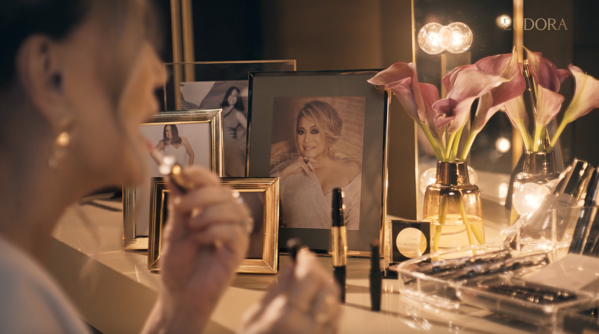 Uma mulher está sentada em uma penteadeira aplicando maquiagem em frente a um espelho.