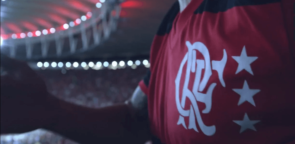 Um homem com a camisa vermelha e preta do Flamengo está parado em frente a um estádio.