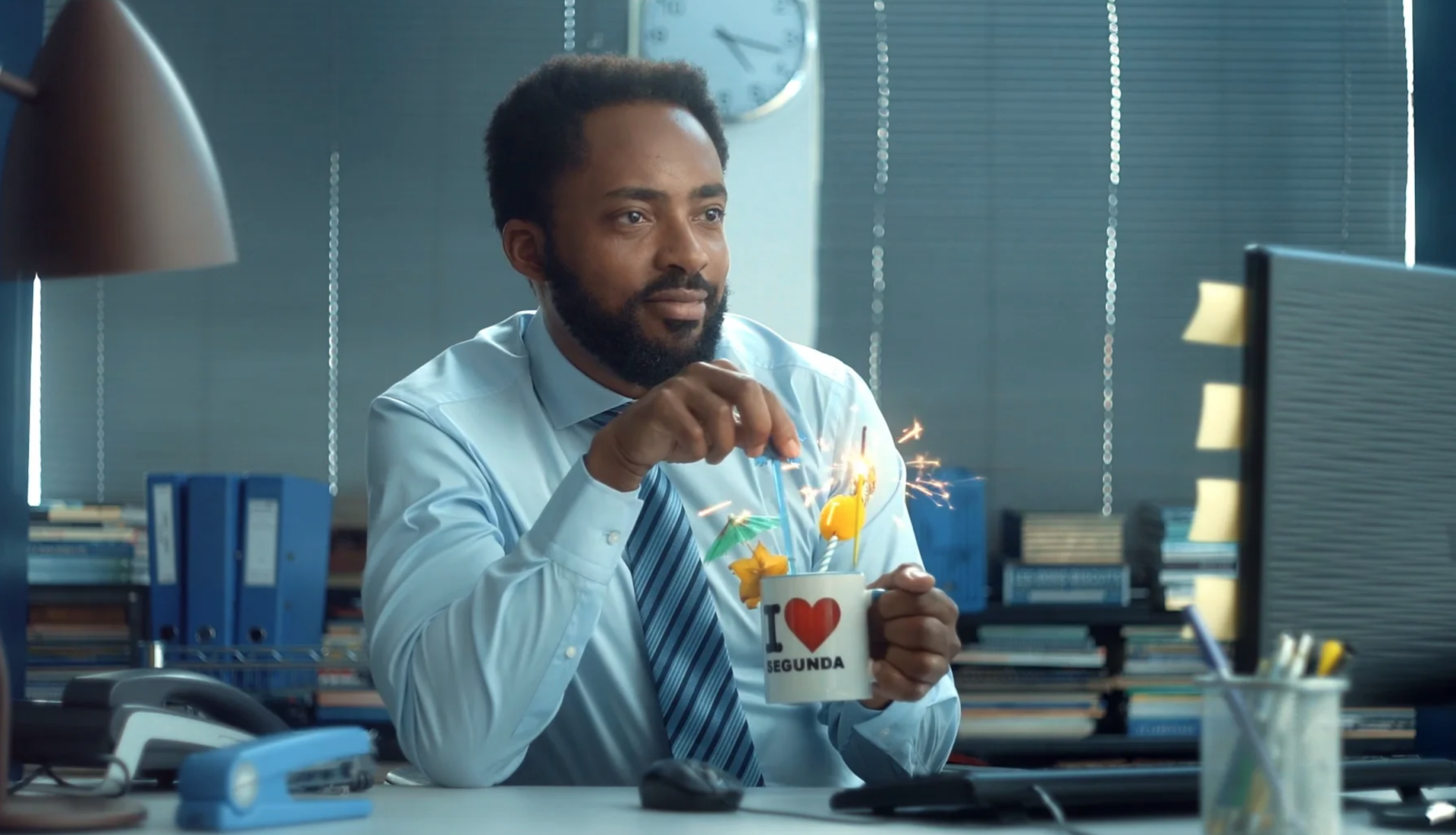 Um homem está sentado em uma mesa em frente a um computador segurando uma xícara de café.
