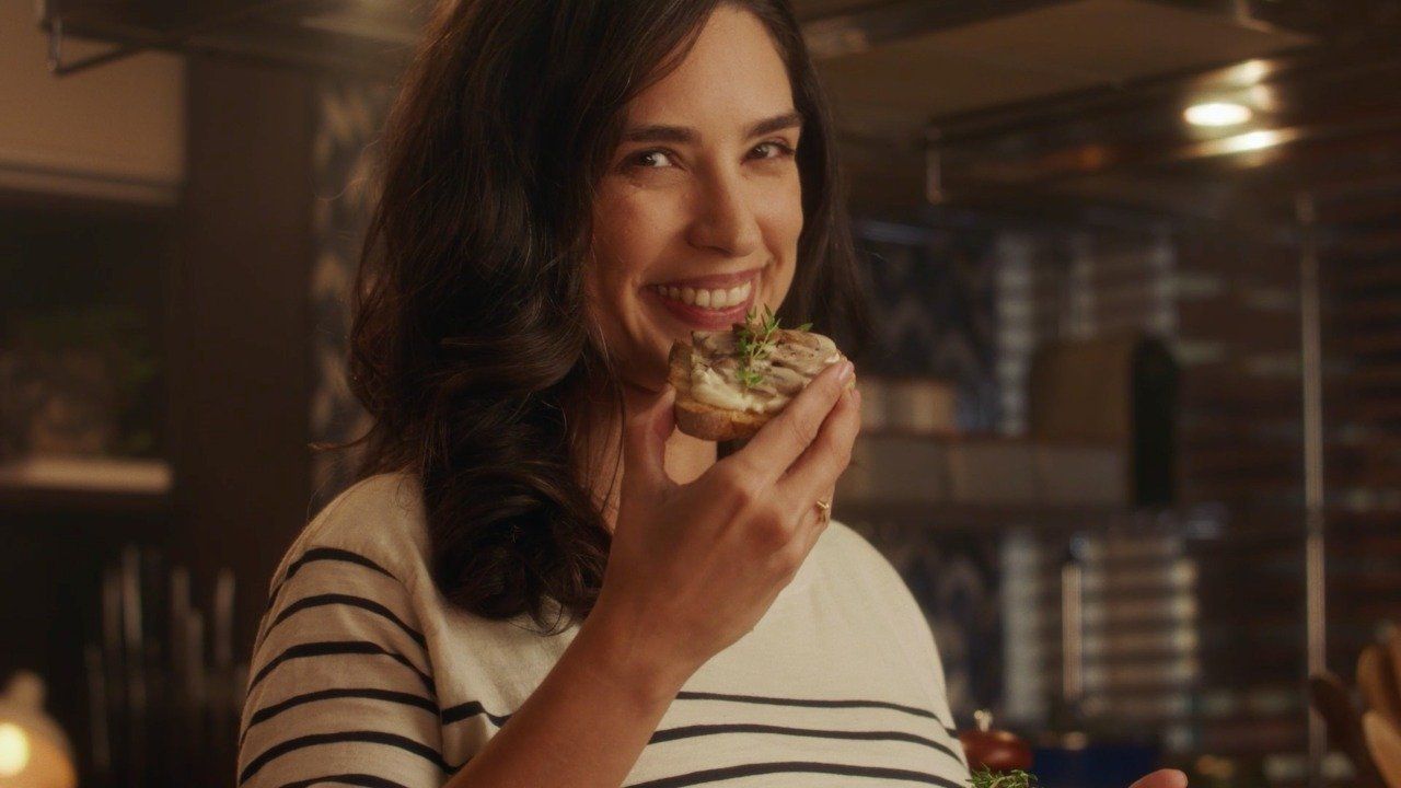 Uma mulher está comendo um sanduíche na cozinha e sorrindo.