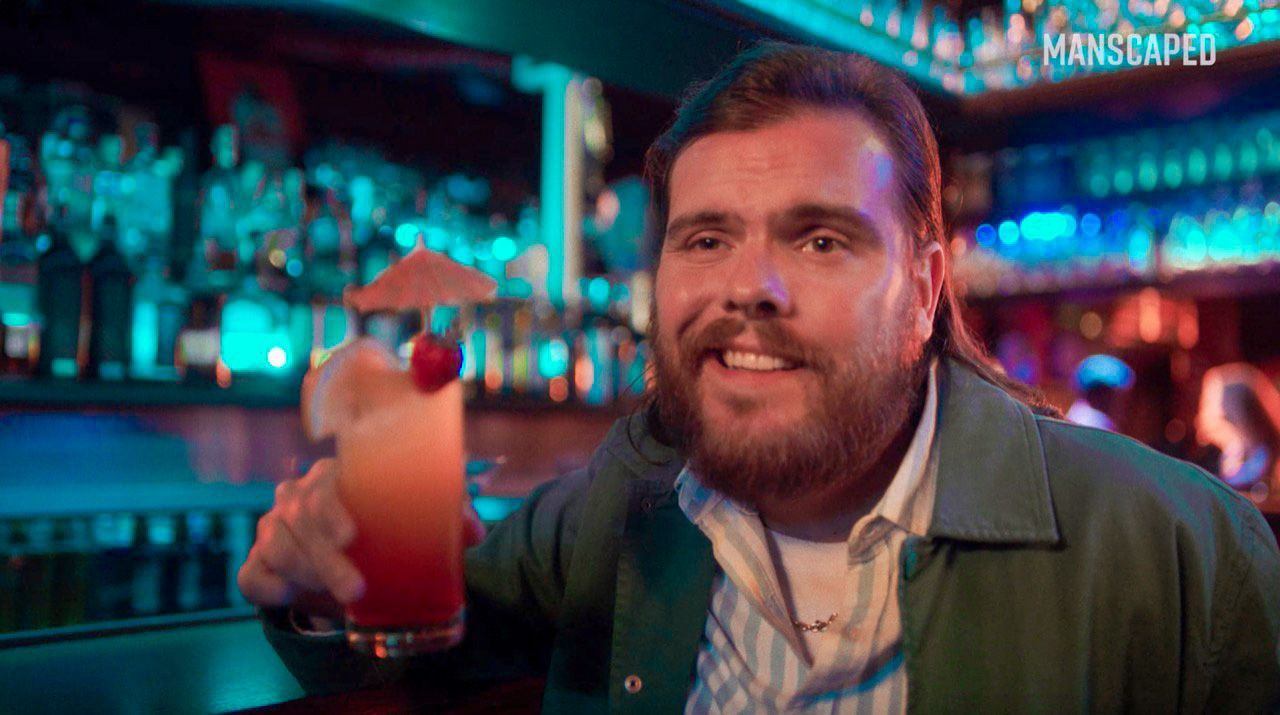 Um homem barbudo está sentado em um bar segurando uma bebida.