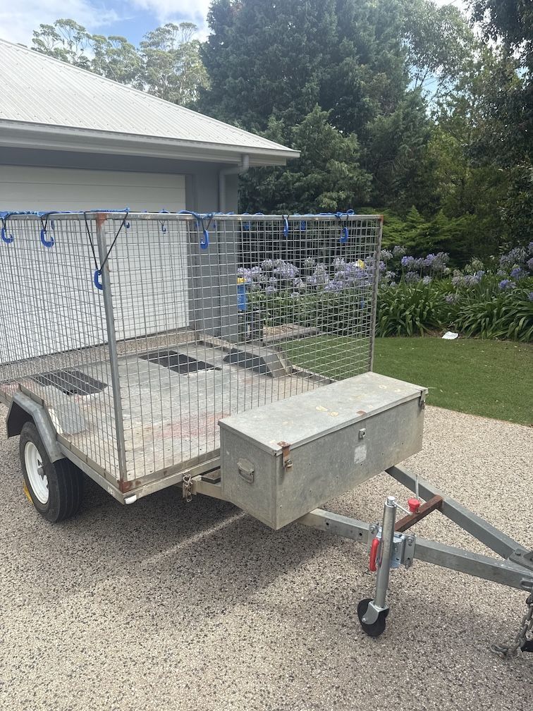 A metal utility trailer with a mesh cage, a front storage box, and a jockey wheel, parked on a paved residential driveway — Resolute Home Services In Toowoomba City, QLD