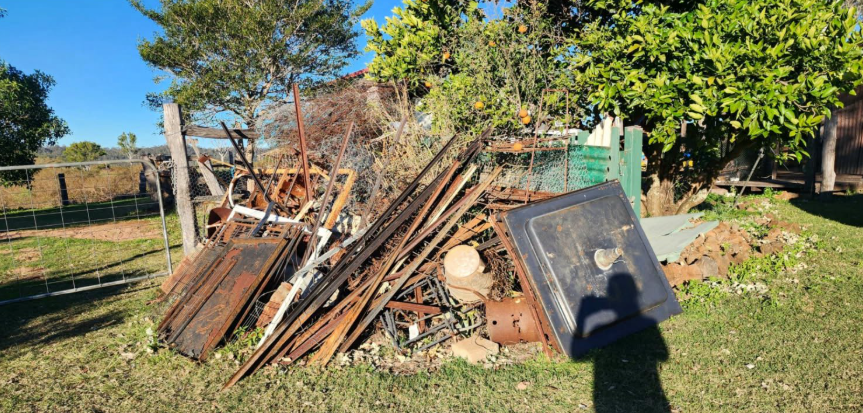 A pile of rusty metal and wood scraps on a grassy lawn with a blue sky. A shadow of a person is in the foreground.