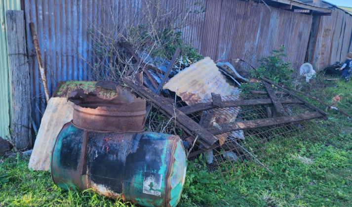 Rusty metal barrels and debris in front of a corrugated metal wall, outdoors.