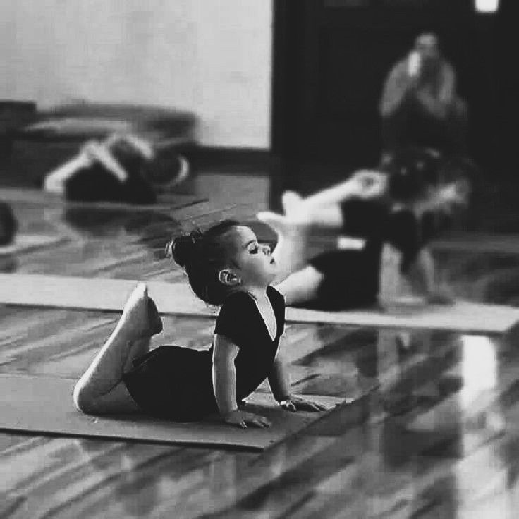 Young girl in black leotard doing yoga on a mat, with other people blurred in the background.