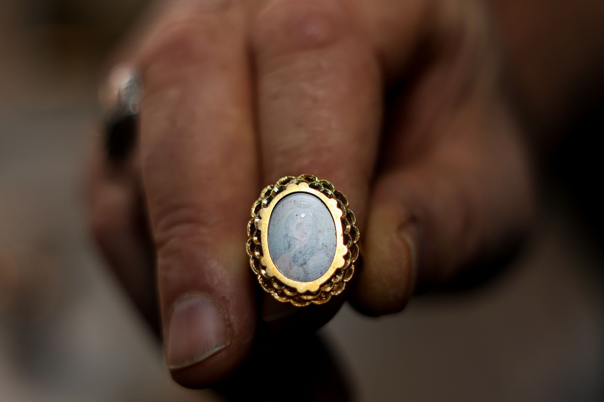 Close-up of a person holding a gold ring with an oval, light-colored stone and ornate design.
