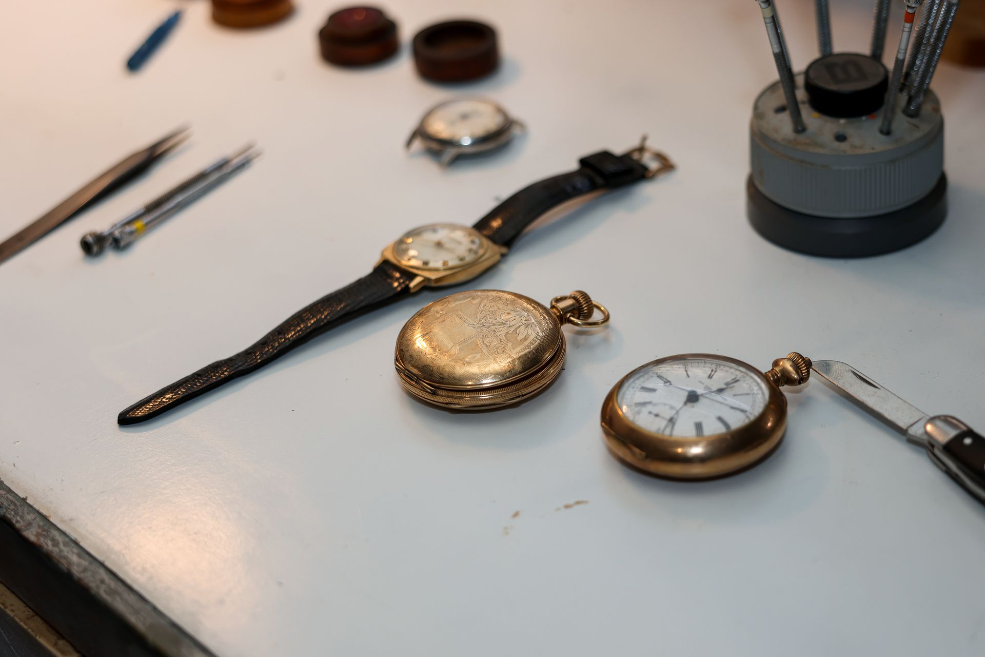 Watches and tools on a white workbench: a wristwatch, two pocket watches, and watchmaking instruments.