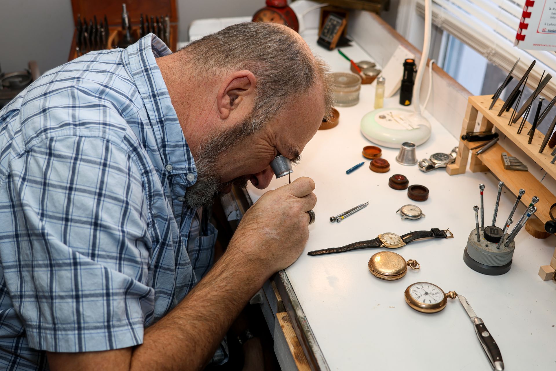 Watchmaker using a loupe to examine a watch on a workbench with tools and parts.