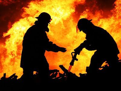 Two firefighters are standing in front of a large fire.