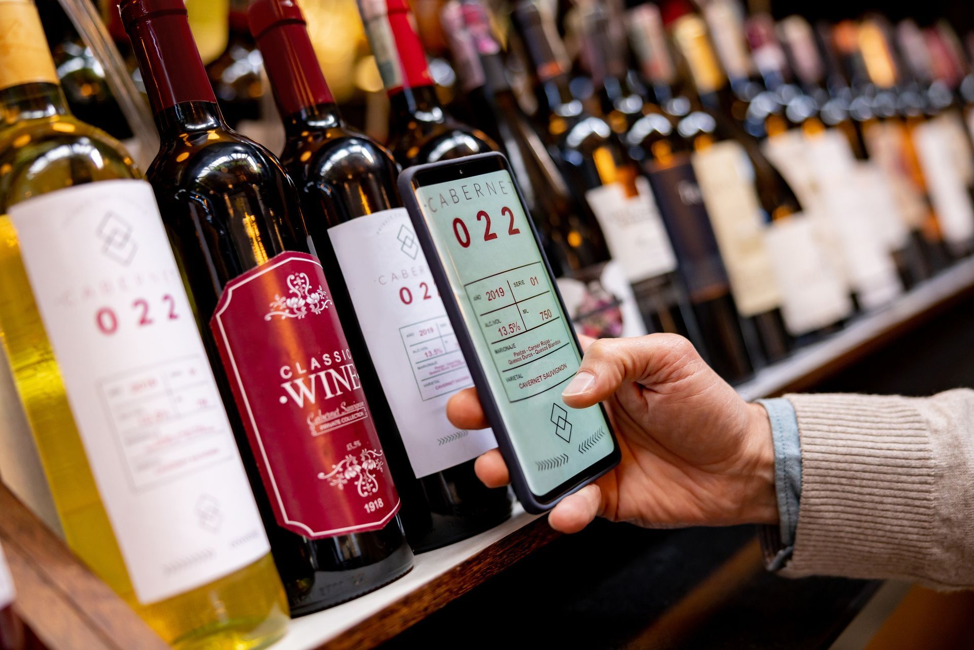A person is holding a cell phone in front of a shelf of wine bottles.