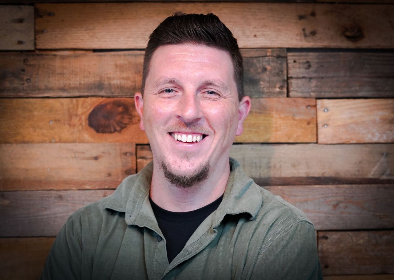 A man in a green shirt is smiling in front of a wooden wall.