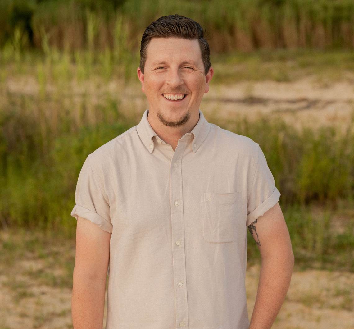 A man in a green shirt is smiling in front of a wooden wall.