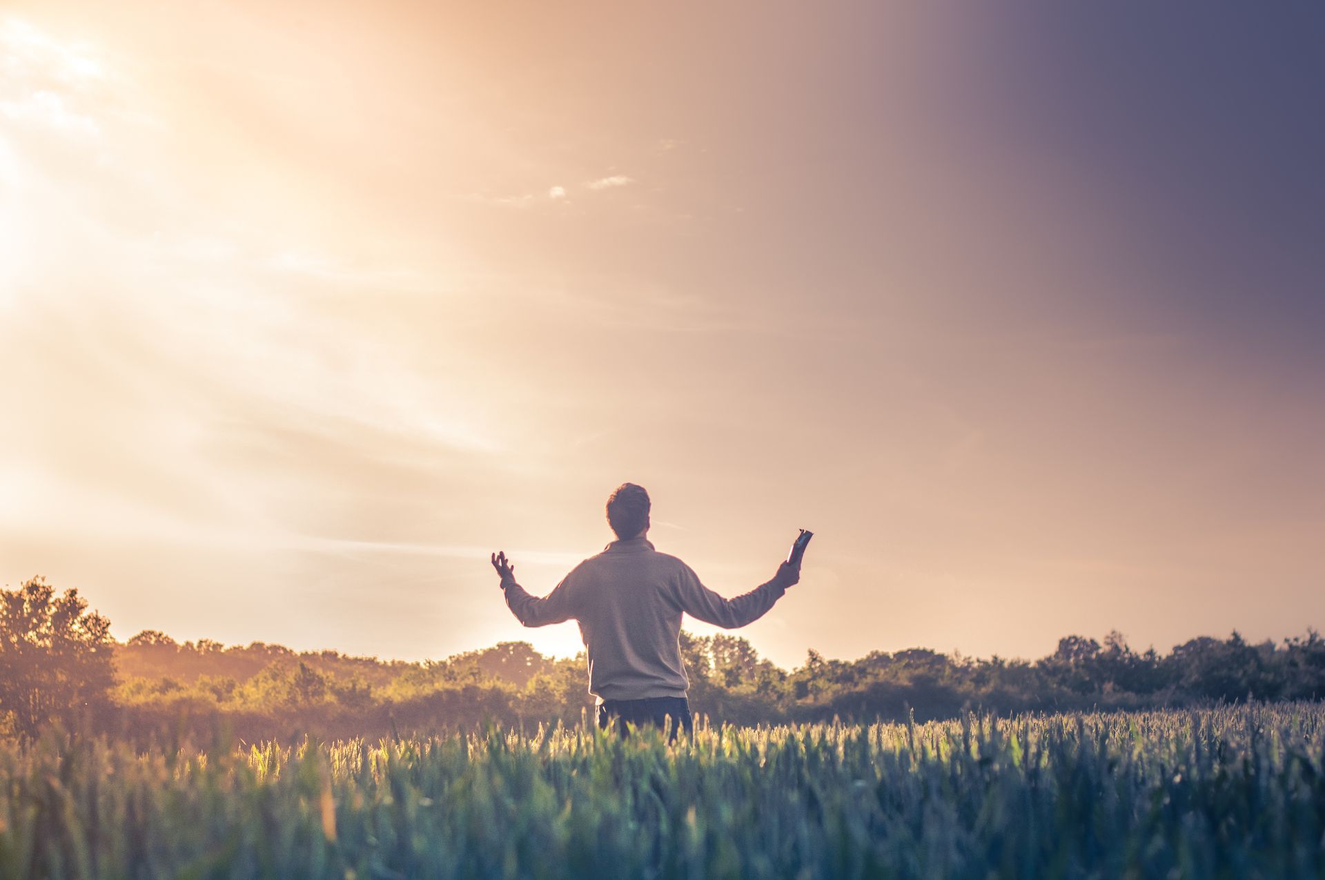 A man is standing in a field with his arms outstretched at sunset.