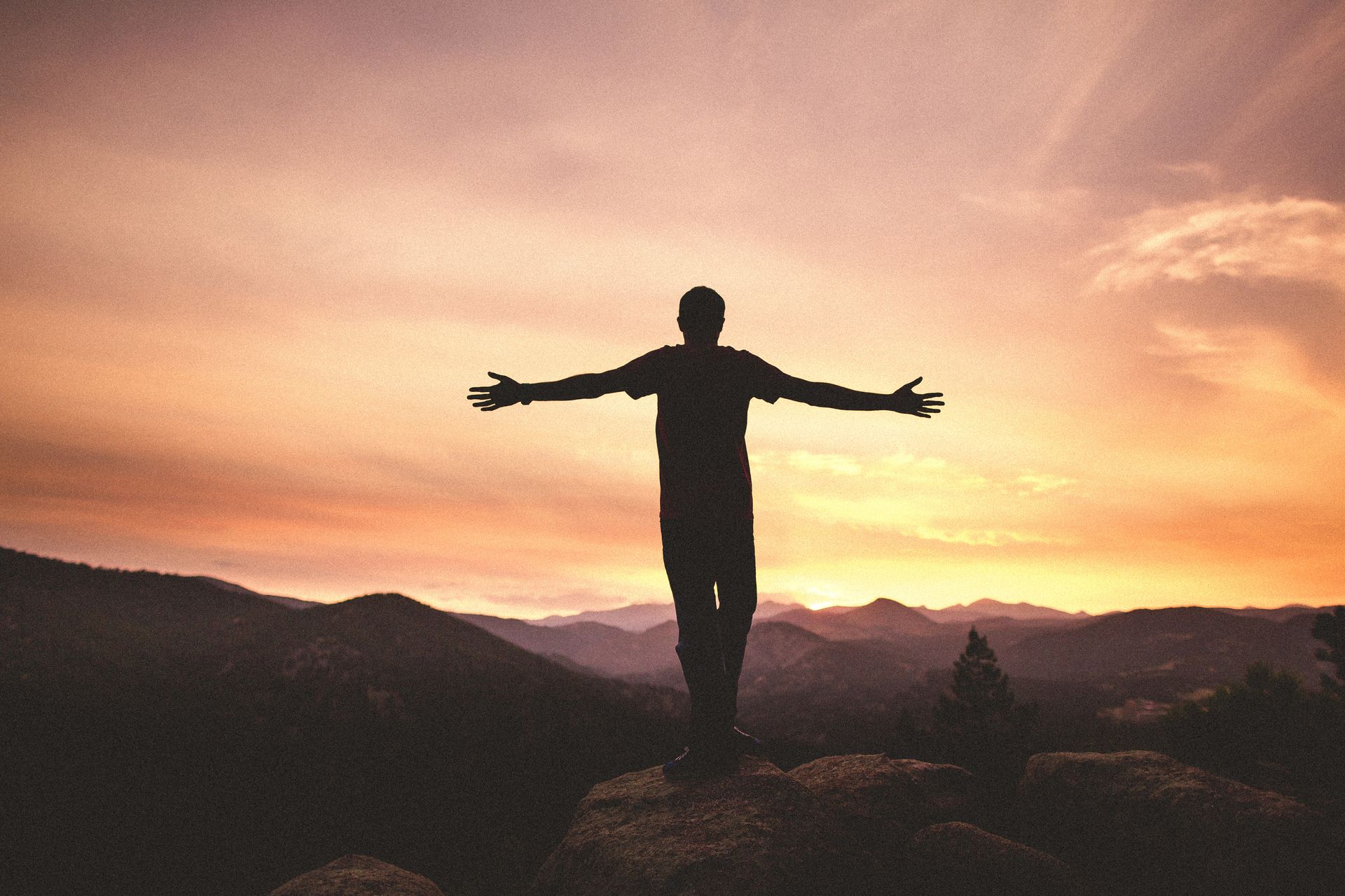 A man is standing on top of a mountain with his arms outstretched at sunset.