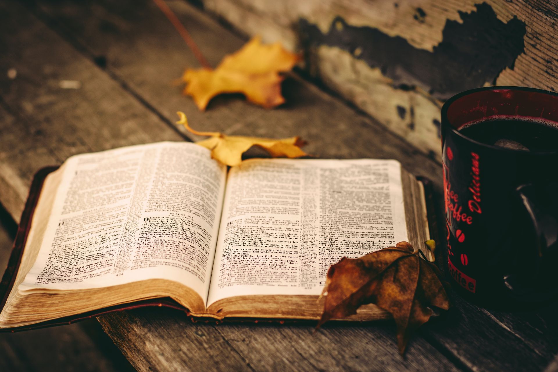 Open Bible, fall leaves, coffee mug on rustic wooden surface.
