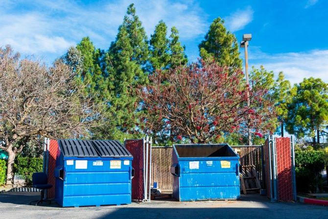 Two blue dumpsters in front of a gated fence, with trees and a clear blue sky behind them.