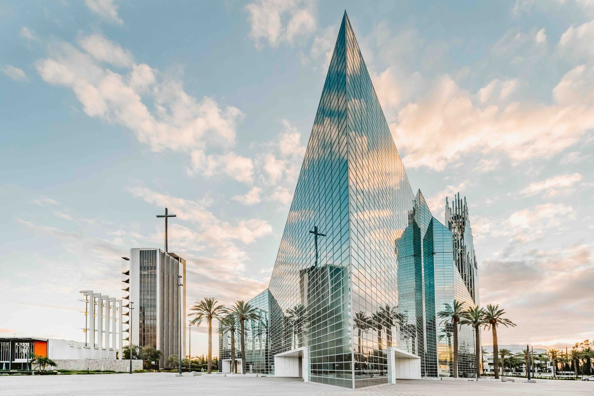Glass-clad church building with a large spire and cross, palm trees, and a bright sky.
