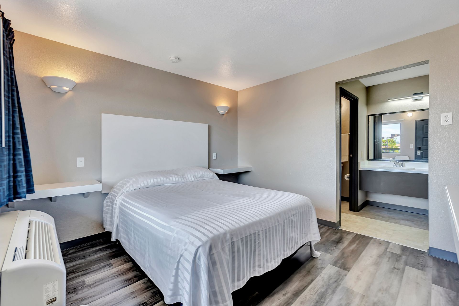 Hotel room with a bed, bathroom in the background, gray walls, and wood-look flooring.