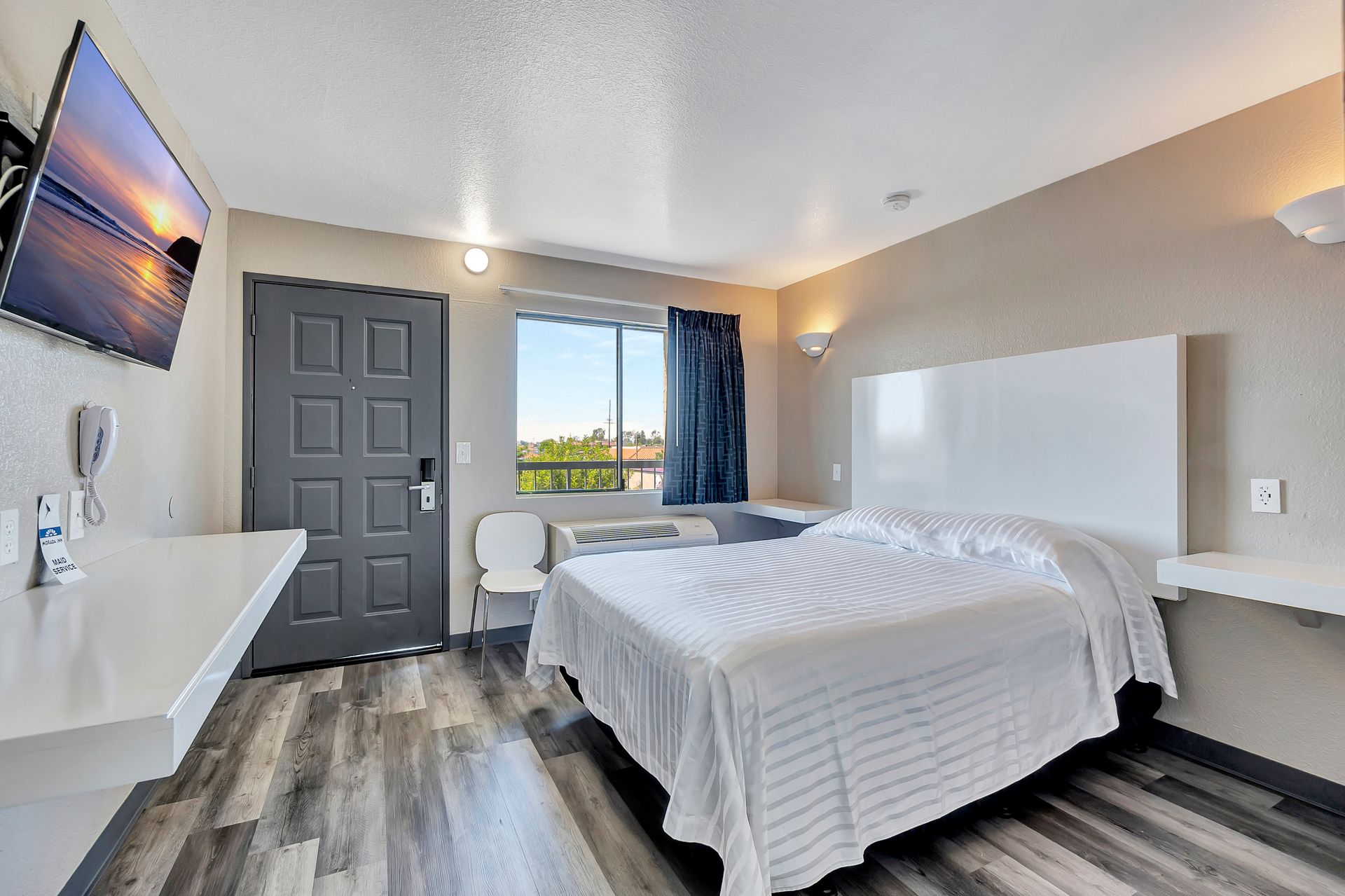 Motel room with a bed, TV, window, and wood-look flooring. Neutral tones.