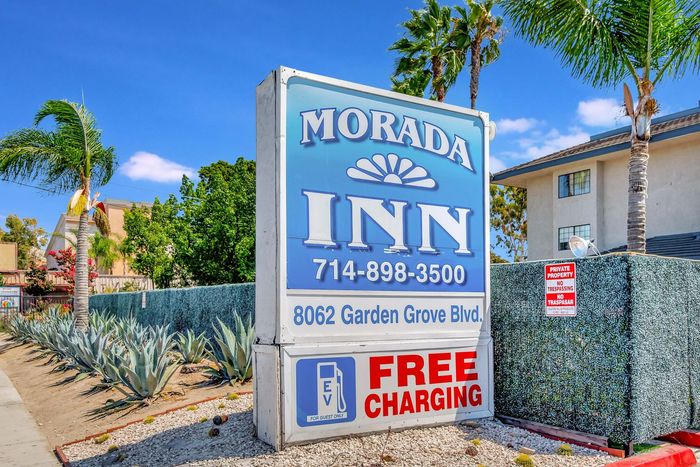 Morada Inn sign with phone number, address, and free charging. Blue sky, palm trees, and fence.