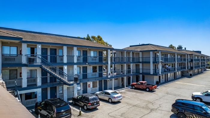 Two-story motel with blue doors, gray siding, and parked cars in a large lot under a blue sky.