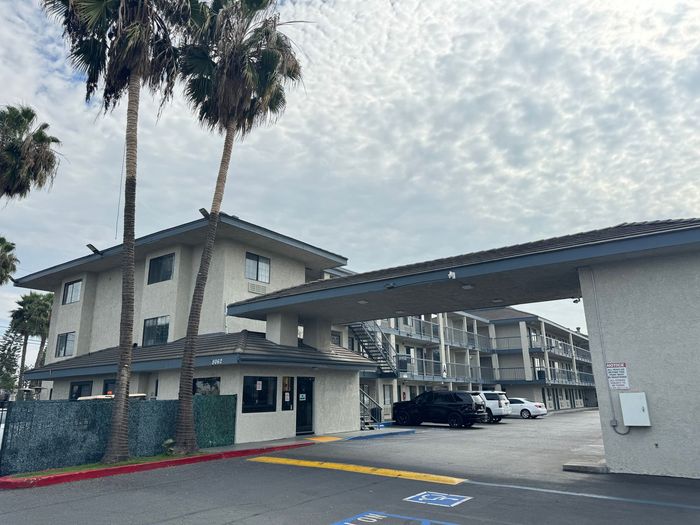 Motel entrance under cloudy sky, palm trees. White buildings, black car, and a security booth.
