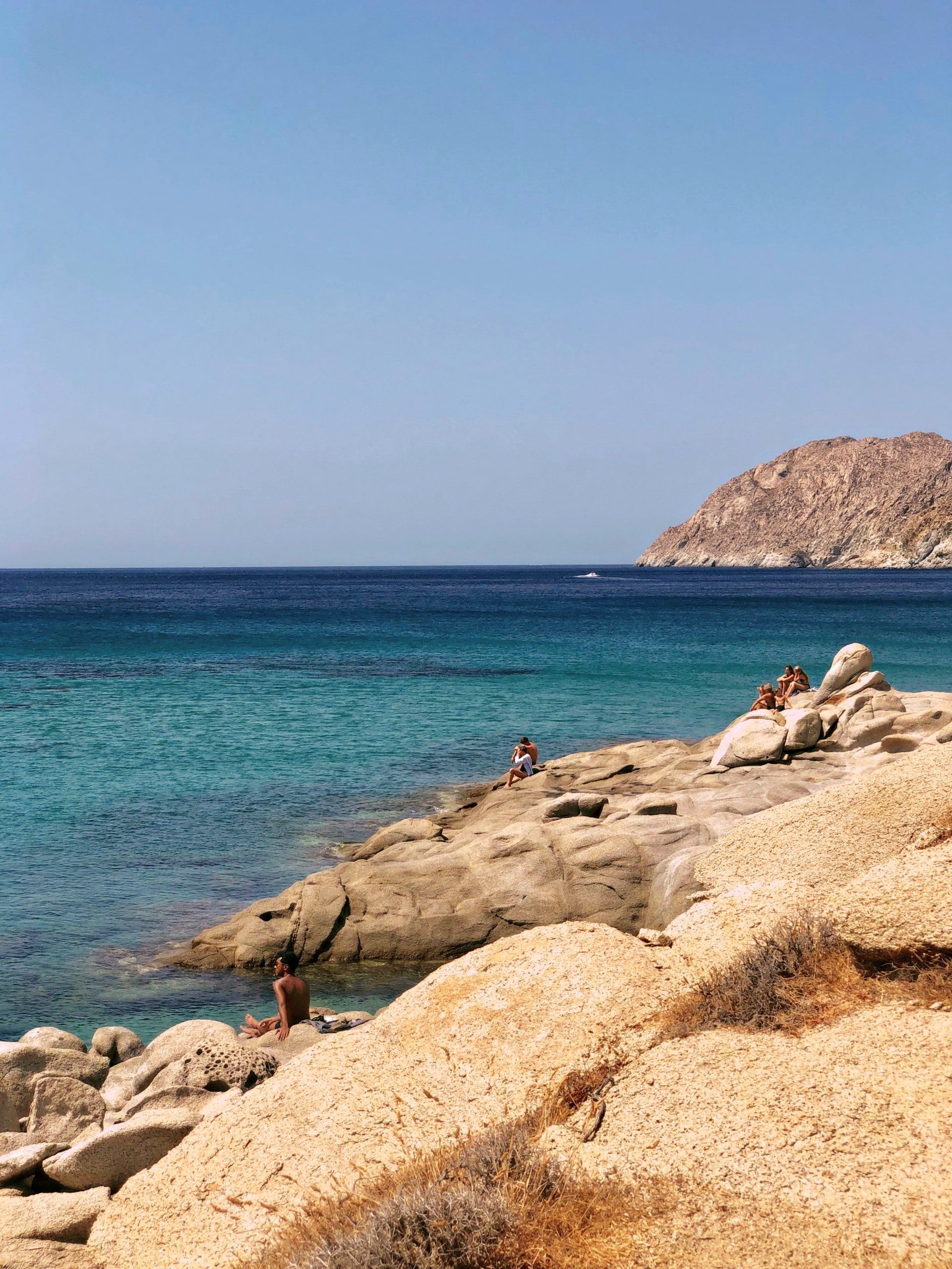 Rocky beach with turquoise water, dark blue sea, and a clear blue sky. People sit on the rocks.