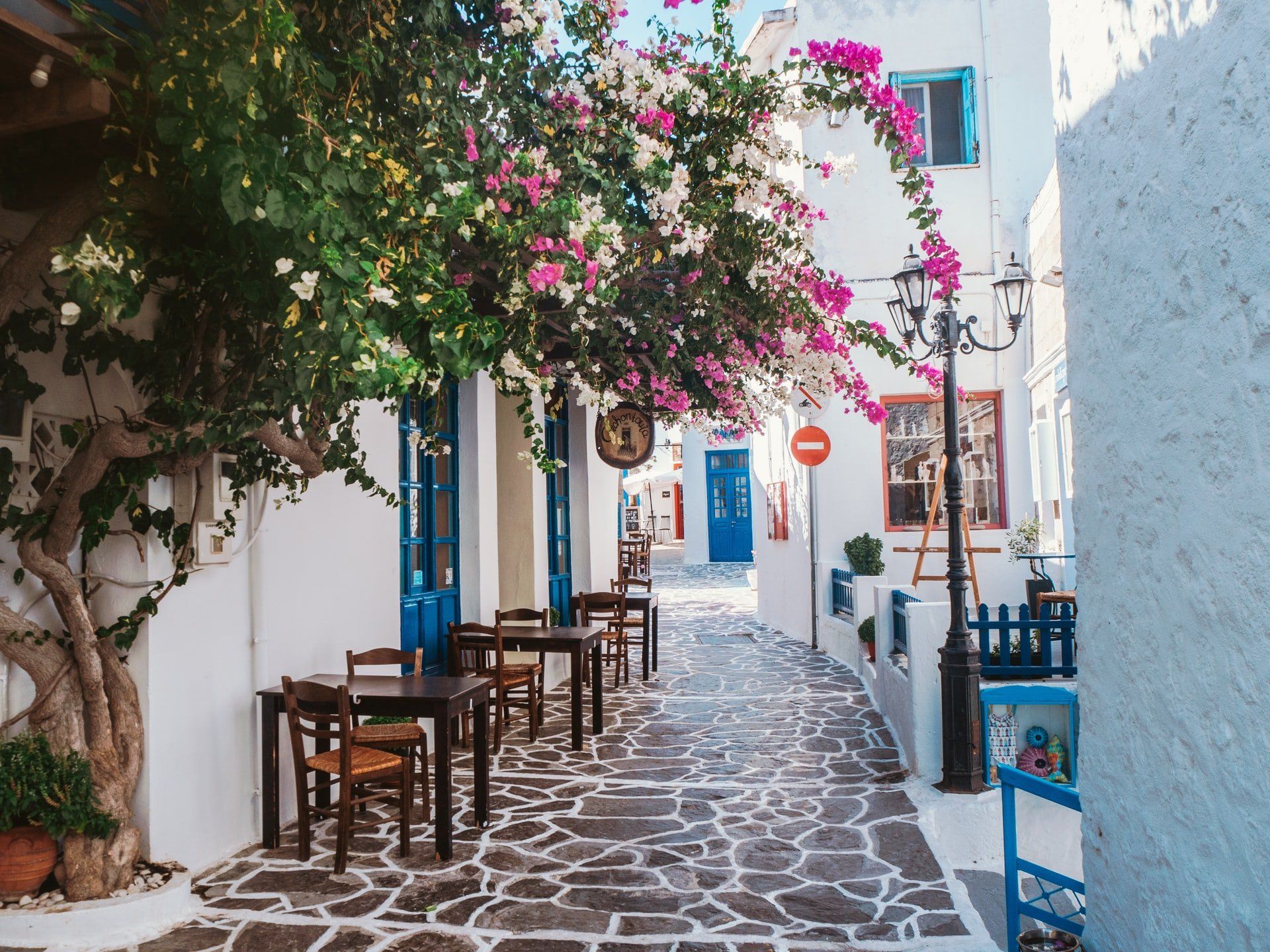Cobblestone street lined with white buildings, blue doors, and tables under a flowering tree in a Mediterranean town.