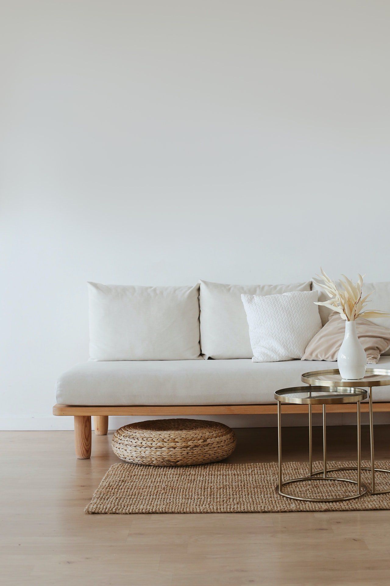 Beige sofa with pillows, two gold side tables, vase, round ottoman, and woven rug against a white wall.