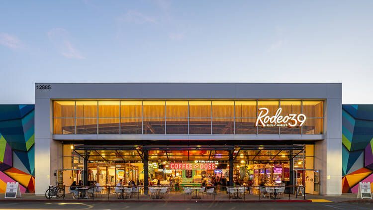 Rodeo 39 Public Market exterior at dusk; people dining and storefronts visible under a bright awning.
