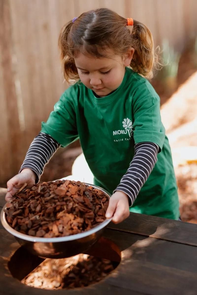 A Little Girl In A Green Shirt Is Holding A Bowl Of Mulch — Moruya Drive Child Care Centre In Port Macquarie, NSW