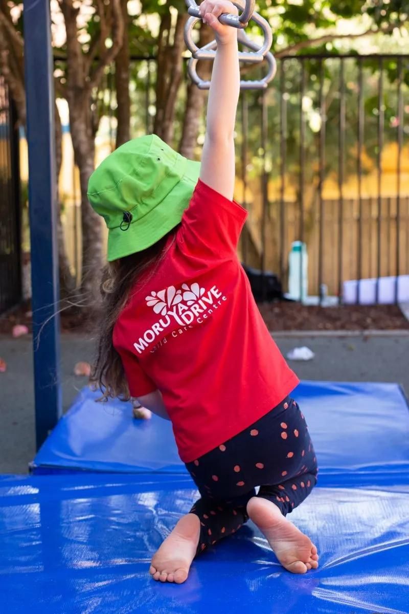 A Little Girl Wearing A Green Hat And A Red Shirt Is Hanging On A Monkey bars — Moruya Drive Child Care Centre In Port Macquarie, NSW