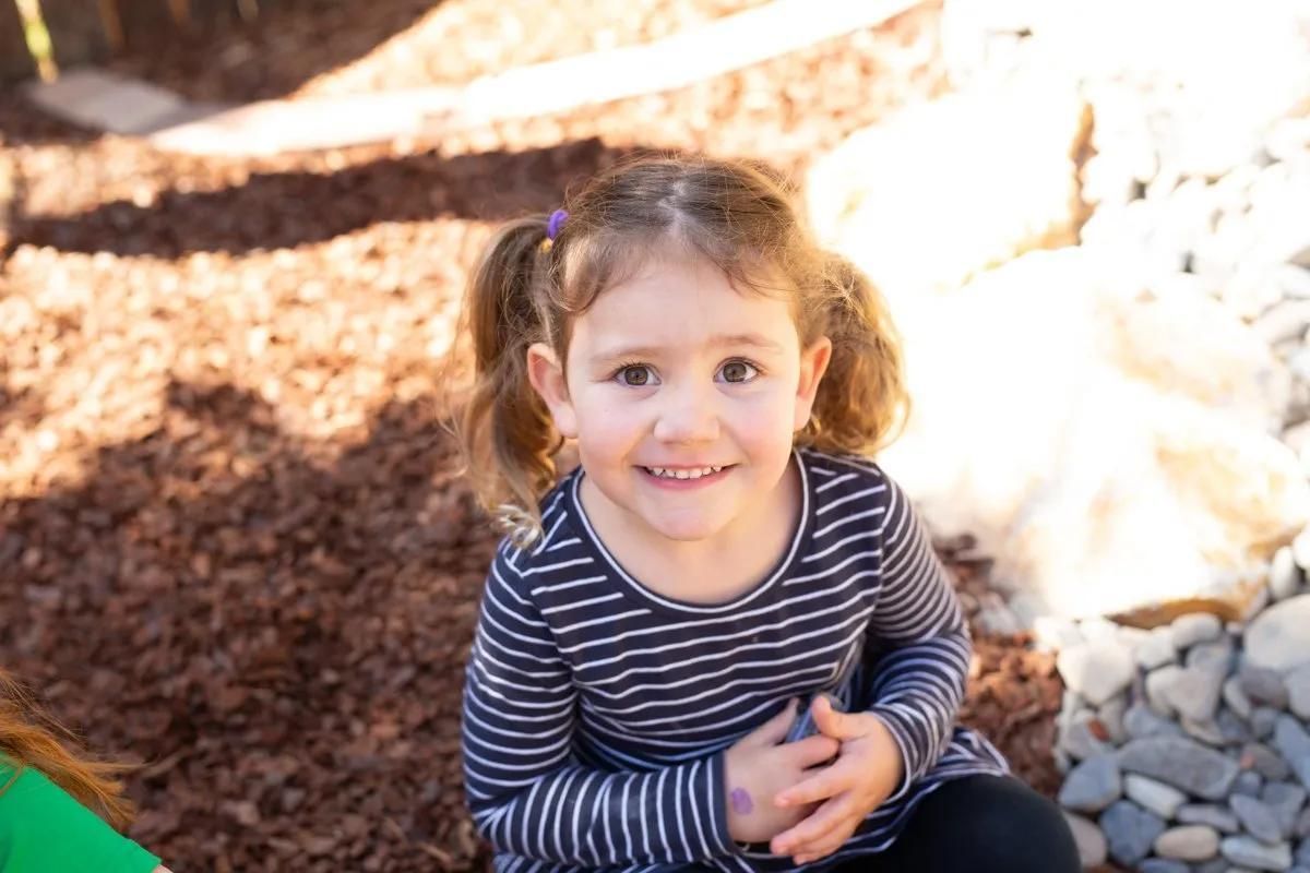 A Little Girl Is Sitting On A Pile Of Mulch And Smiling — Moruya Drive Child Care Centre In Port Macquarie, NSW