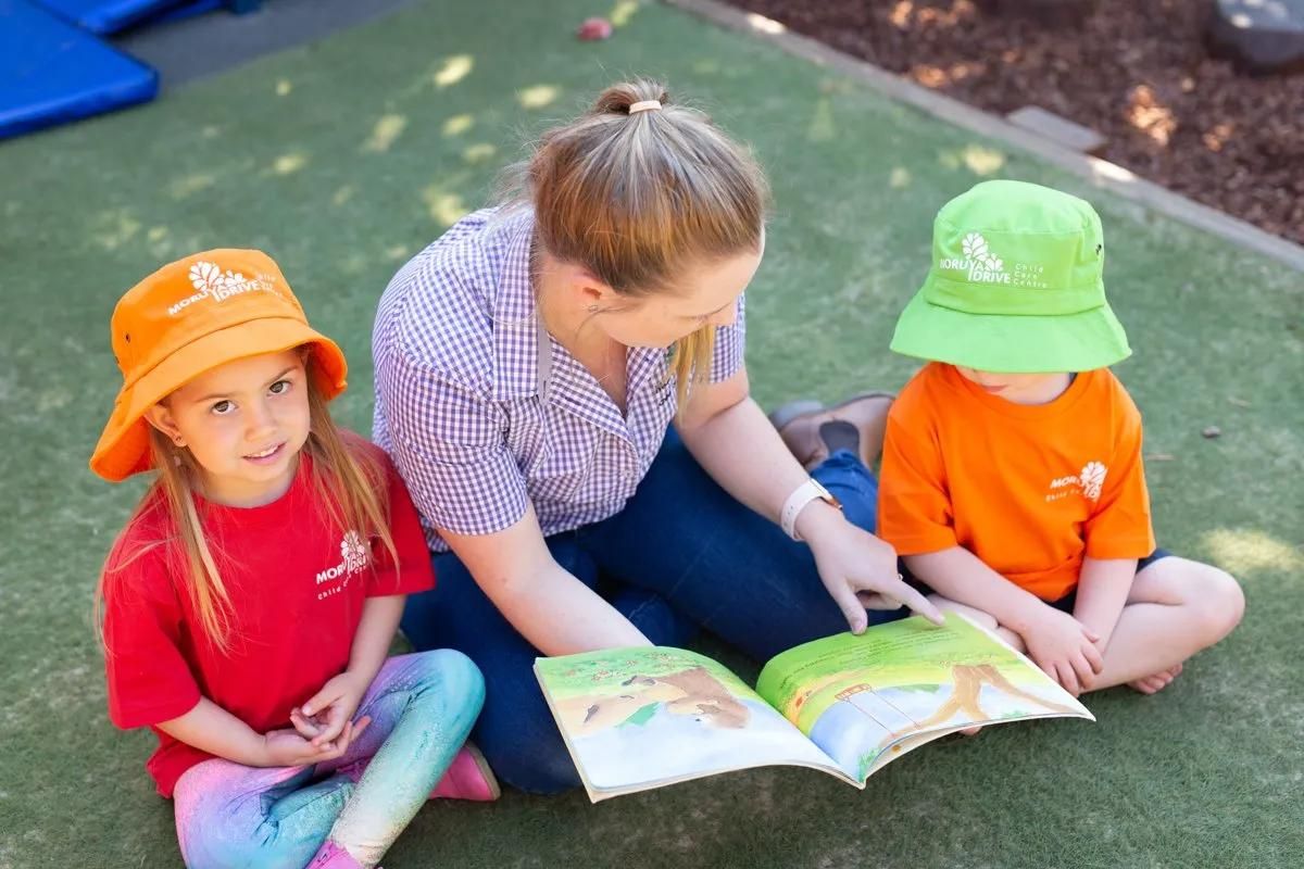 A Woman Is Sitting On The Grass Reading A Book To Two Children — Moruya Drive Child Care Centre In Port Macquarie, NSW