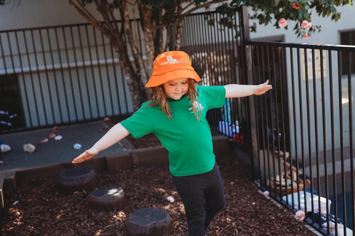 A Young Girl In A Green Shirt And Orange Hat Is Standing On A Rock With Her Arms Outstretched — Moruya Drive Child Care Centre In Port Macquarie, NSW