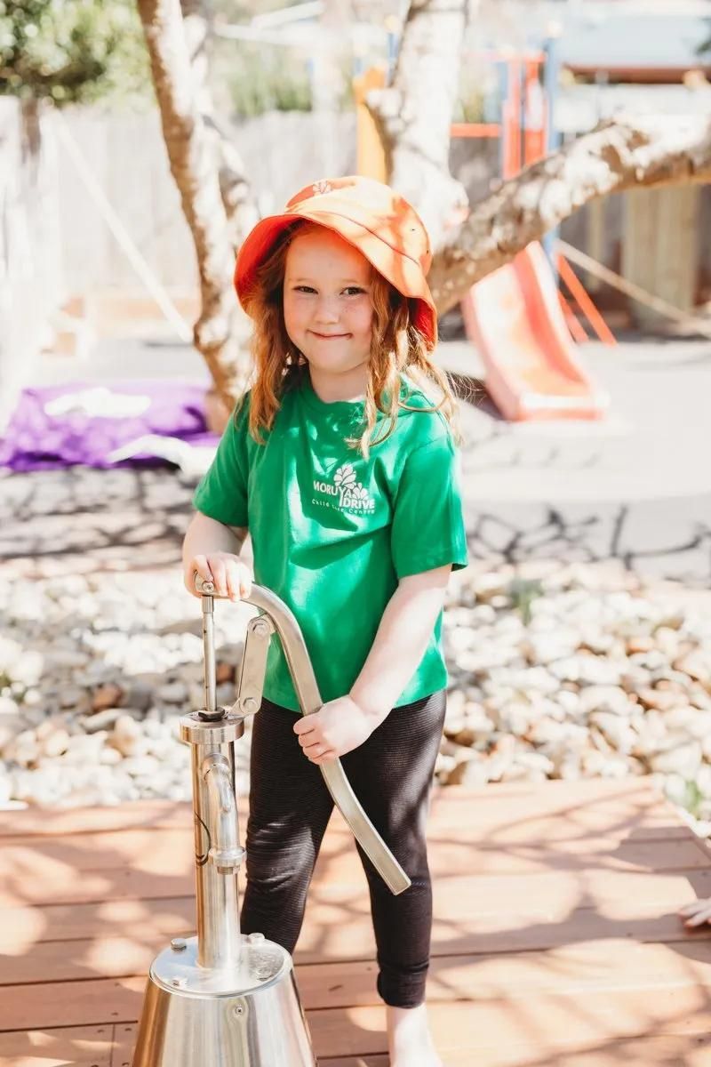 A Little Girl In A Green Shirt And Orange Hat Is Playing With A Water Pump — Moruya Drive Child Care Centre In Port Macquarie, NSW