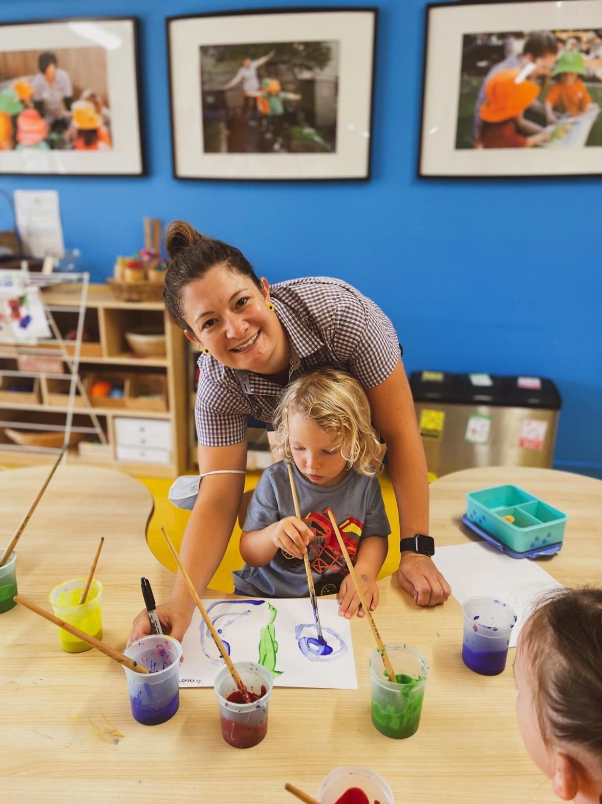 A Woman Is Teaching A Child How To Paint At A Table — Moruya Drive Child Care Centre In Port Macquarie, NSW
