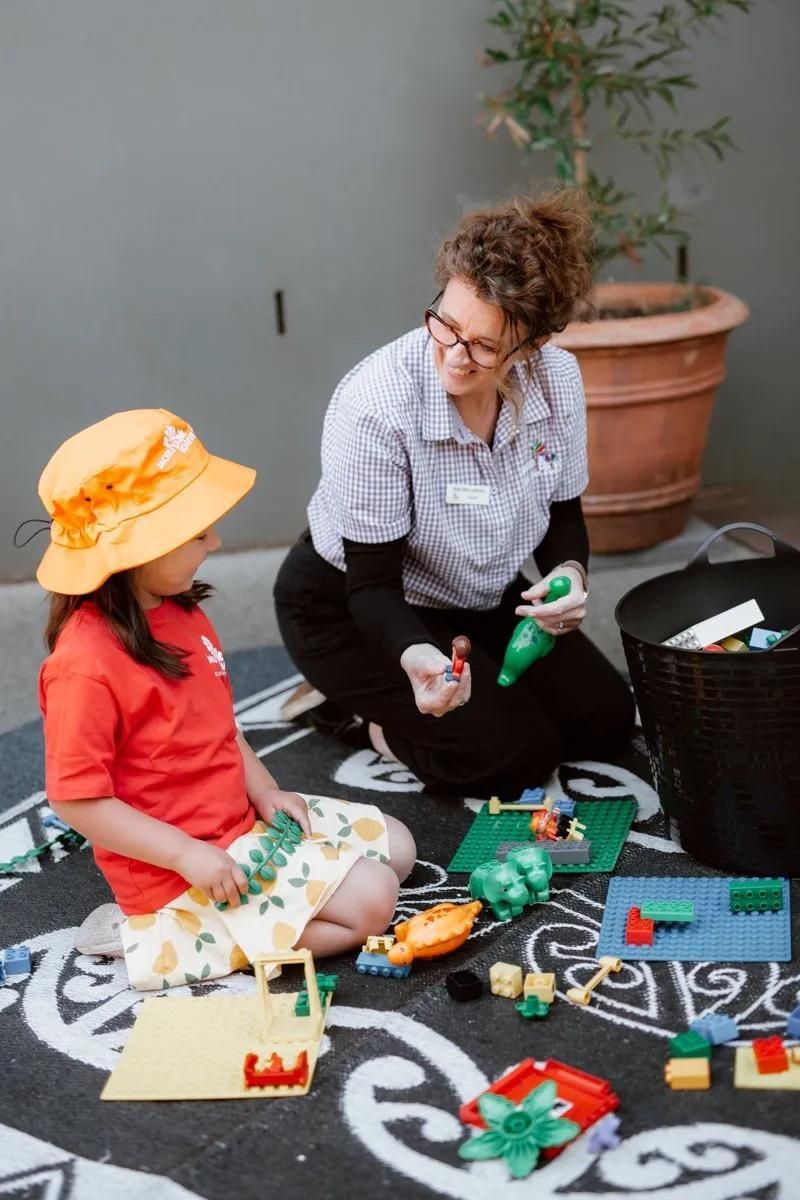 A Woman Is Kneeling Down Next To A Little Girl Playing With Toys — Moruya Drive Child Care Centre In Port Macquarie, NSW