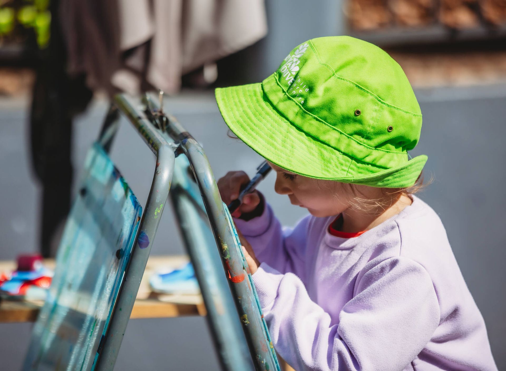 A little girl wearing a green hat is painting on an easel — Moruya Drive Child Care Centre In Port Macquarie, NSW