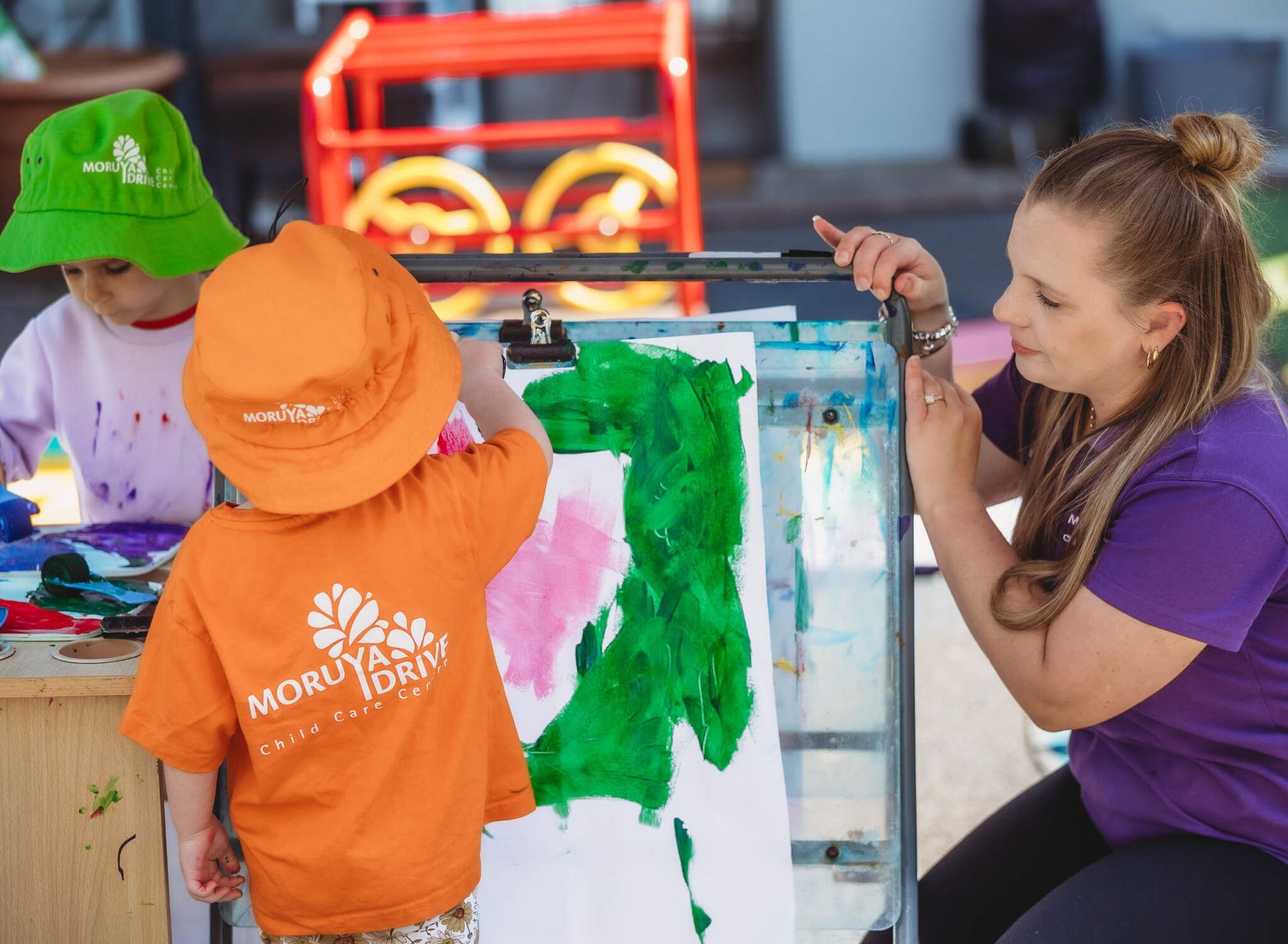 A woman is teaching a child how to paint on a canvas — Moruya Drive Child Care Centre In Port Macquarie, NSW
