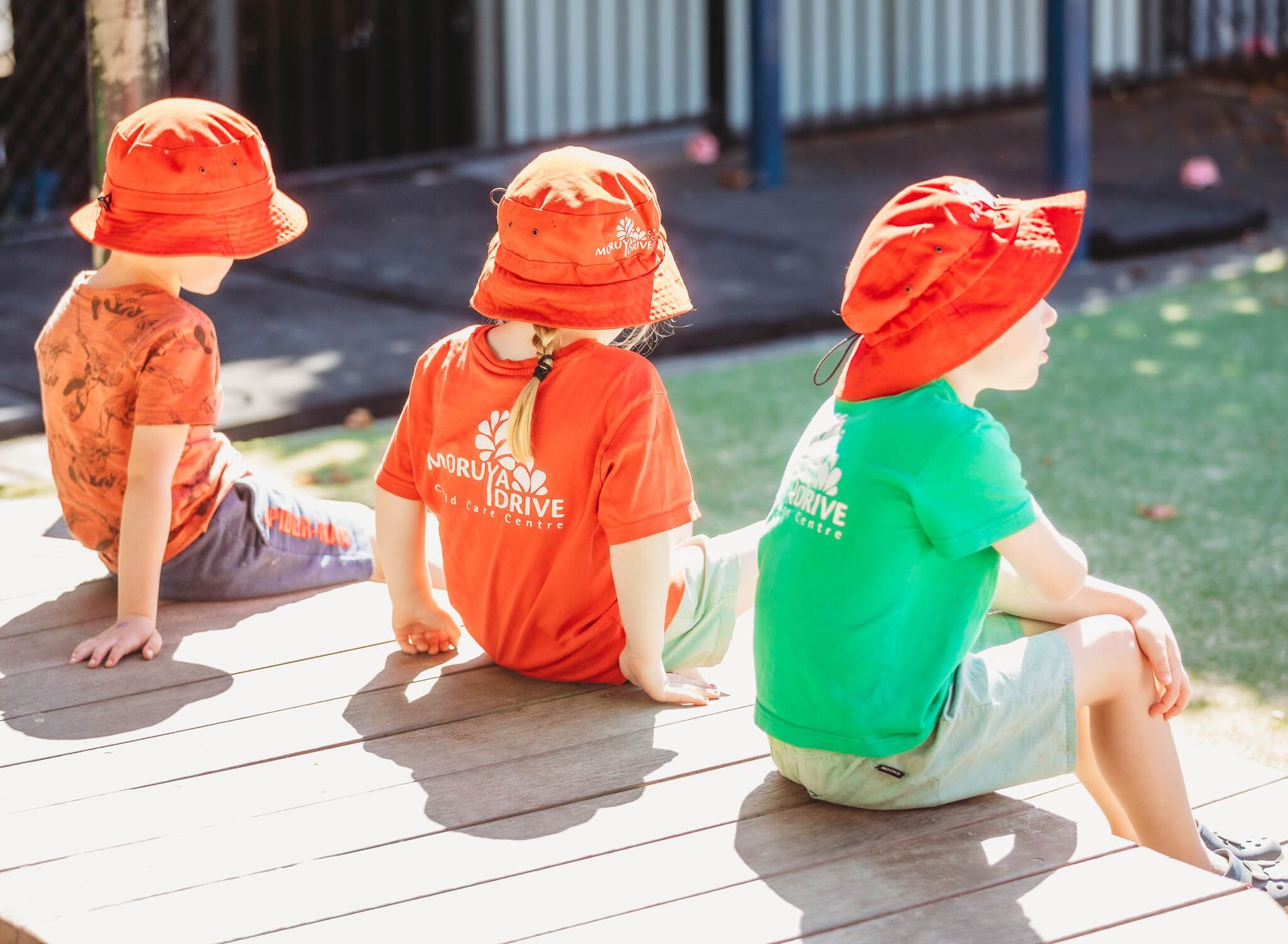 Three children are sitting on a wooden deck wearing red hats — Moruya Drive Child Care Centre In Port Macquarie, NSW
