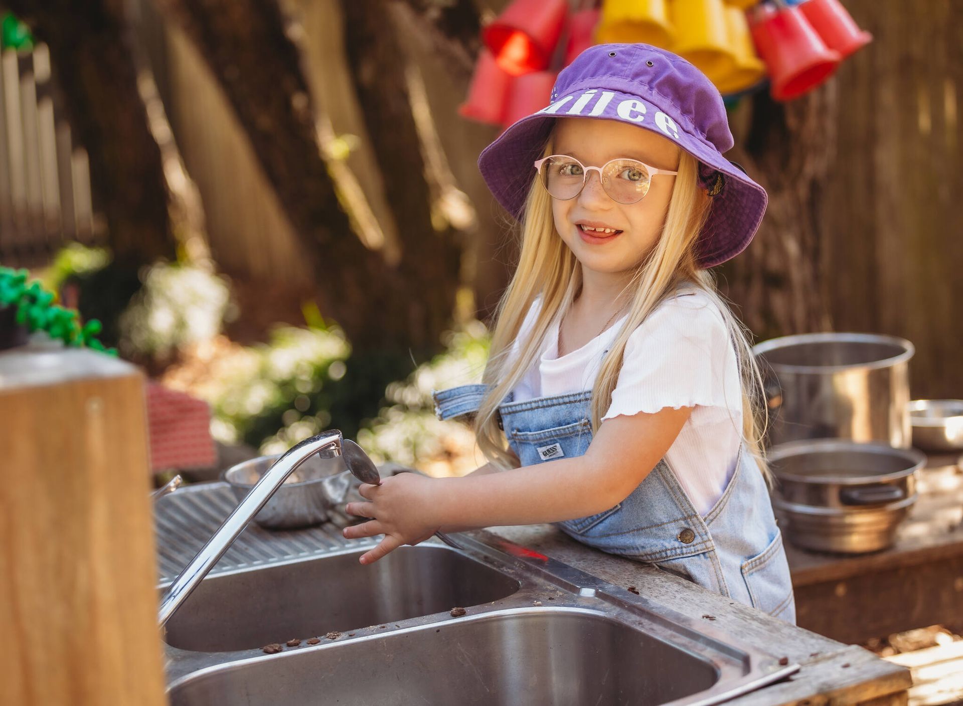 A little girl wearing a purple hat and glasses is washing dishes in a sink — Moruya Drive Child Care Centre In Port Macquarie, NSW