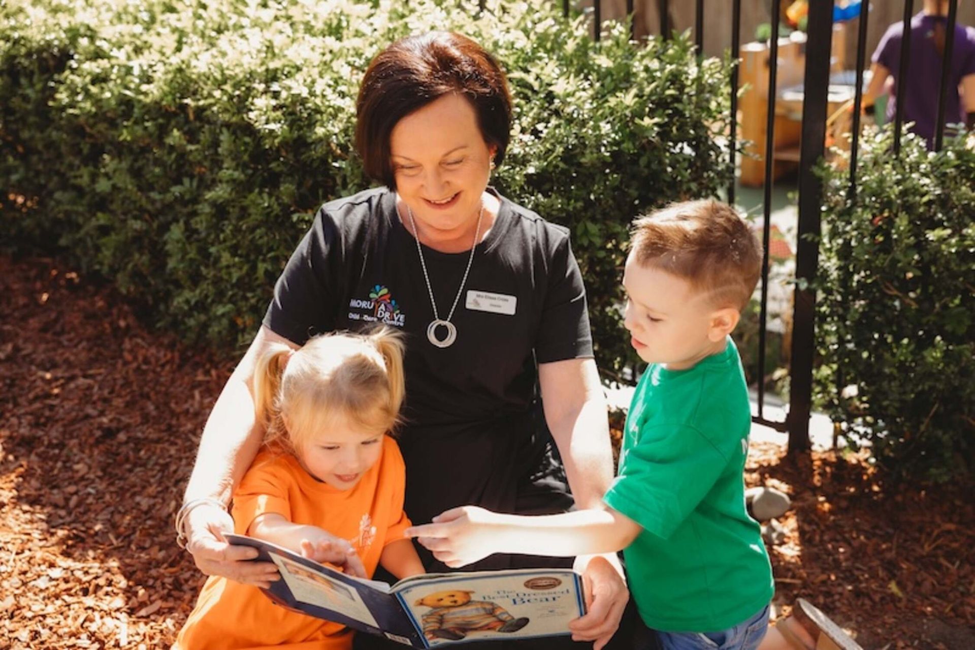 A woman is reading a book to two children — Moruya Drive Child Care Centre In Port Macquarie, NSW