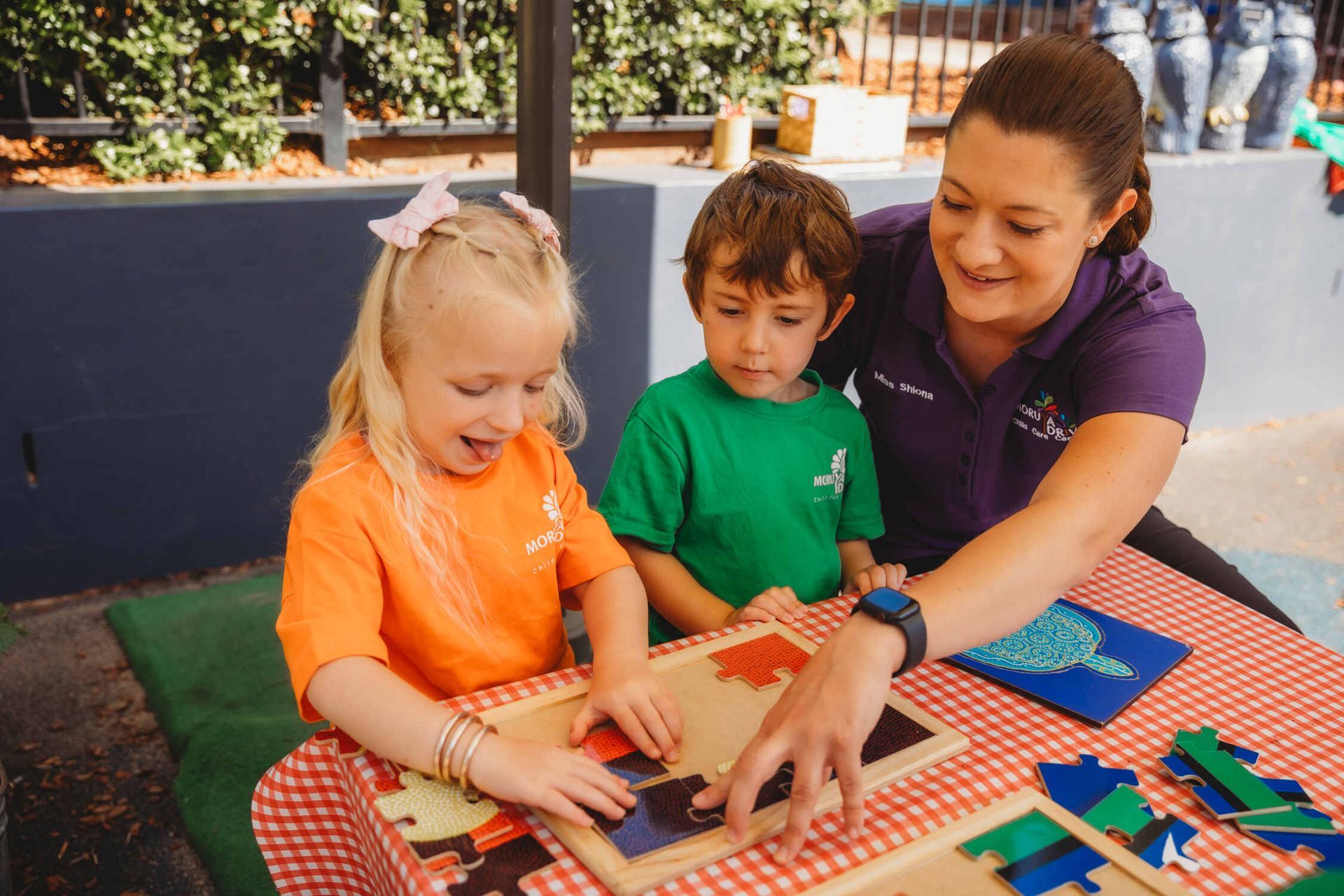 A woman is sitting at a table with two children playing with puzzles — Moruya Drive Child Care Centre In Port Macquarie, NSW