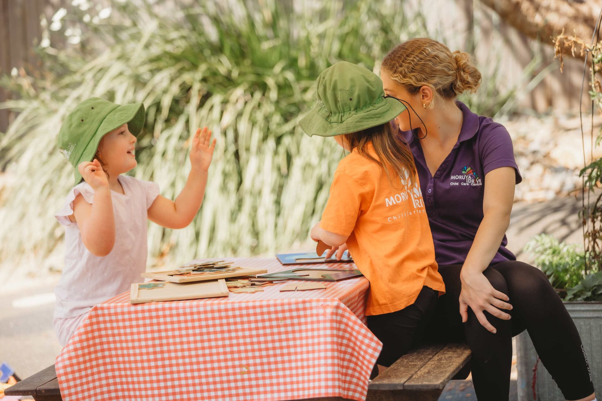 A woman and two children are sitting at a picnic table — Moruya Drive Child Care Centre In Port Macquarie, NSW